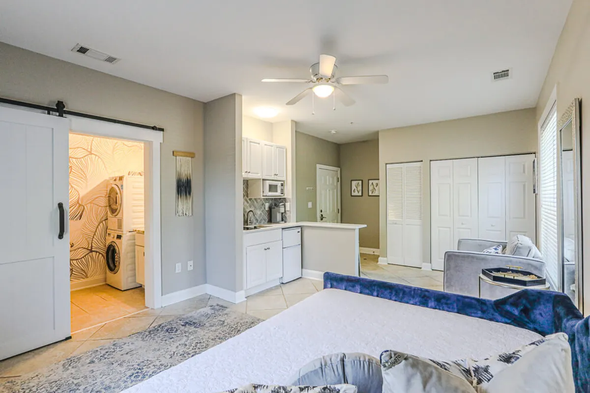 A well-lit studio main room combining a bed, kitchenette, seating area, and laundry zone. Modern furnishings create functional zones for relaxing, cooking, socializing, and doing laundry, all woven together in an open plan. Daylight streams through large windows, illuminating the cozy, versatile space.