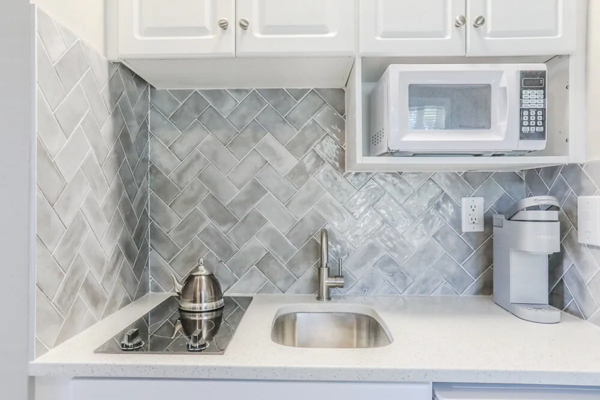 This image features a compact yet modern kitchenette, highlighted by a striking gray herringbone backsplash, a stainless steel kettle, a microwave, and a coffee maker. The setting is bright and inviting, with a functional layout that makes the most of limited space.
