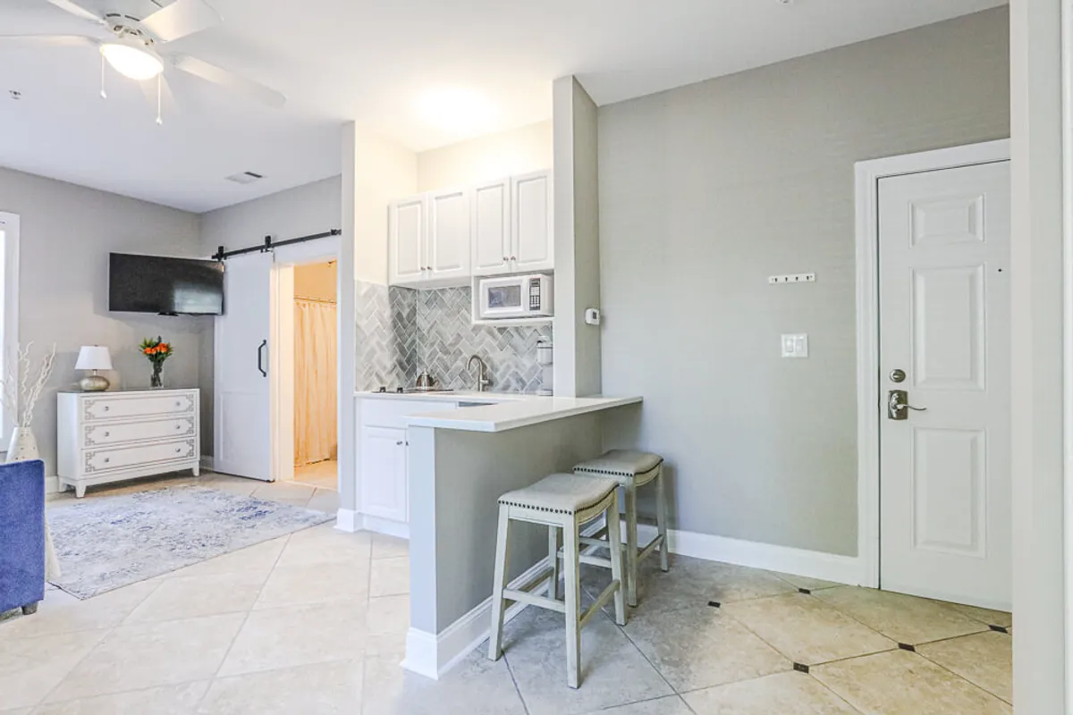 A welcoming kitchenette setup with two bar stools at a breakfast bar, decorative flowers, a television, and a dresser nearby. The space feels comfortable and is designed for casual meals, morning routines, or socializing while cooking.