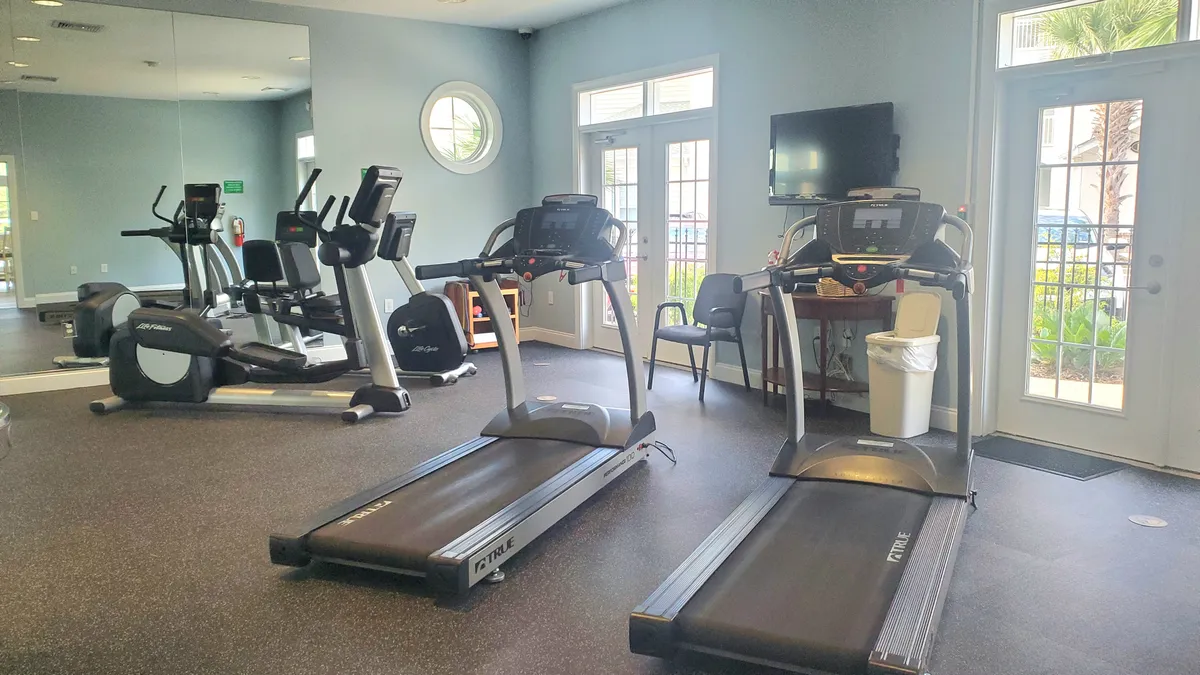 A bright, well-equipped fitness center featuring treadmills and exercise bikes positioned in front of expansive windows, offering a motivating atmosphere for workouts. A television and mirrored wall complete the energetic, communal environment.