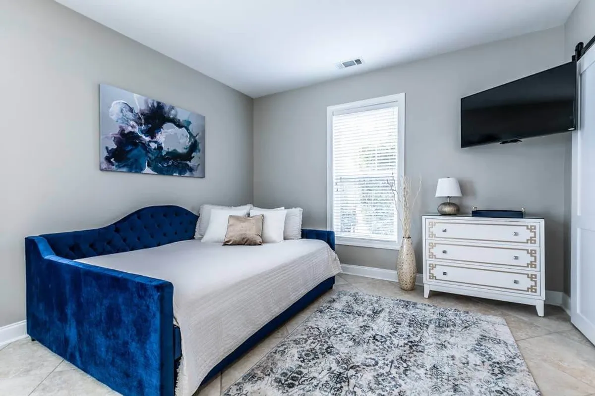 A sunlit, modern studio interior featuring a plush blue velvet daybed positioned beneath abstract art, a sleek white dresser, and a window streaming in natural light. The scene exudes tranquility and invites guests to unwind with a book or enjoy peaceful moments by the window.