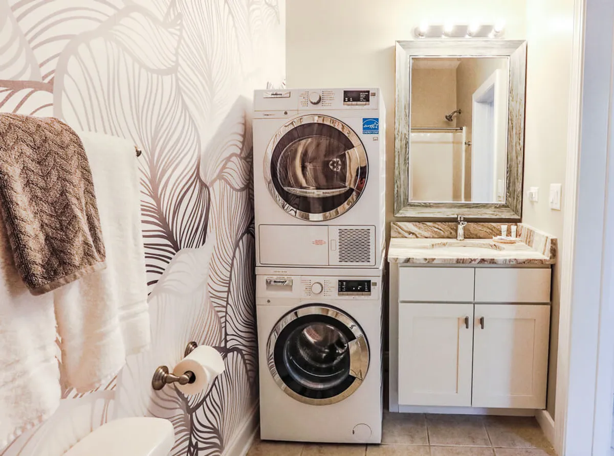 A pristine interior scene with a stacked washer and dryer, luxurious marble countertop, decorative mirror, and neatly folded textured towels. The clean lines, quality finishes, and careful organization evoke a sense of order and understated luxury, reinforcing the idea of pampering daily routines.