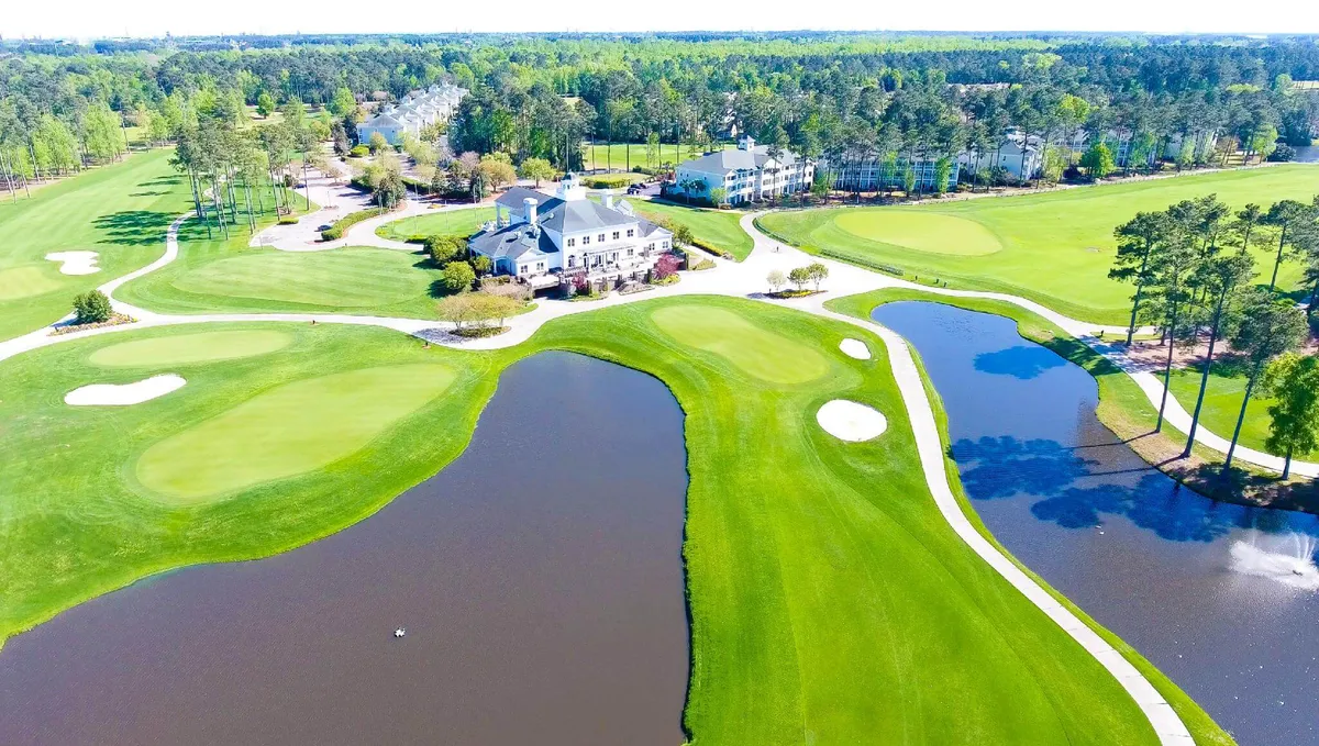 This exterior image captures the lush green expanse of the golf course with the clubhouse prominently in the background. Water features and surrounding mature trees create a tranquil, picturesque setting. The well-maintained fairways and manicured landscape are clearly visible, highlighting the premium golfing environment.