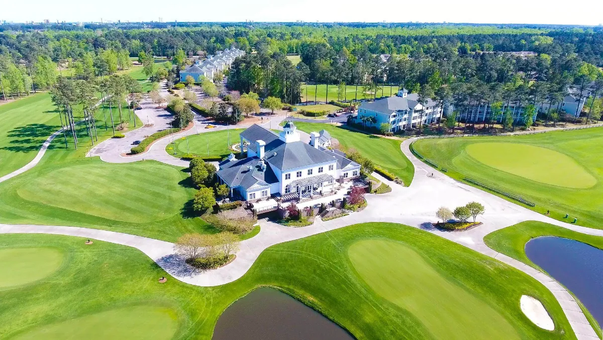 This exterior shot shows the golf clubhouse from a different angle, with manicured lawns, lush golf course greens, shimmering ponds, and mature trees stretching into the distance. The setting sun creates a warm, leisurely ambiance.
