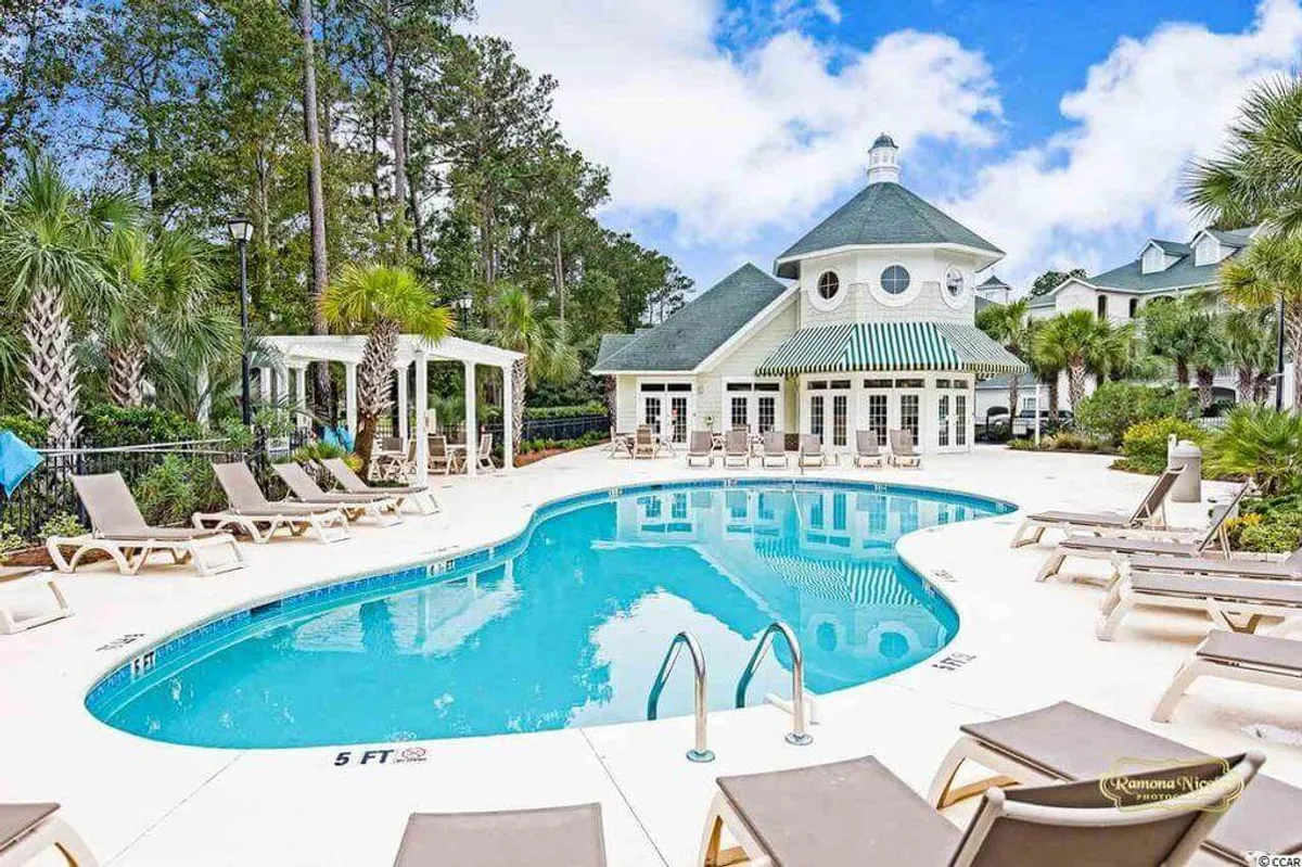An inviting view of the sparkling swimming pool surrounded by lounge chairs, with the elegant clubhouse and swaying palm trees in the background. The scene is bustling with guests relaxing, socializing, and enjoying the tranquil resort atmosphere.