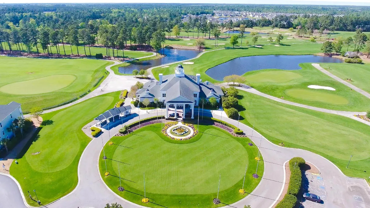 The photo features a wide view of the elegant clubhouse, surrounded by beautifully landscaped gardens and glimpses of the golf course and a scenic pond. The upscale, peaceful atmosphere is emphasized by the open green spaces and inviting clubhouse structure.