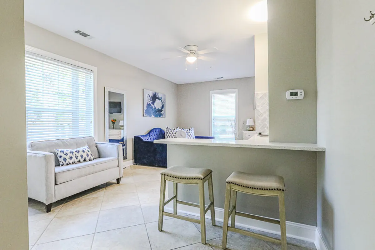 A stylish, inviting clubhouse interior with a plush sofa, bar stools, contemporary artwork, and a sunlit window. The space is designed for casual conversations, socializing, and relaxing in comfort—ideal for unwinding after a day by the pool or a workout.