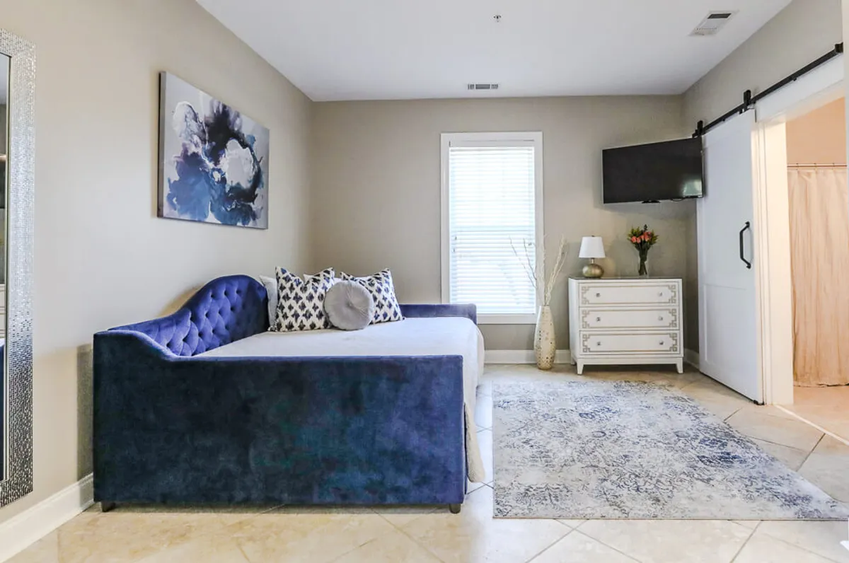 A bright and contemporary living area featuring a comfortable sofa, modern artwork, a decorative lamp, a vibrant floral arrangement, and a patterned rug. Sunlight filters in, creating a cozy and inviting atmosphere ideal for relaxation or family time.