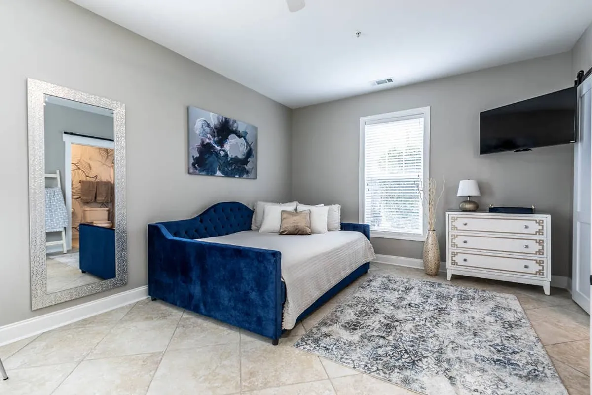 A serene, inviting living area featuring a plush blue velvet daybed, a large mirror that reflects natural light, an abstract painting for artistic flair, and a white dresser with gold accents. The space feels comfortable and restorative, ideal for unwinding after a long day.