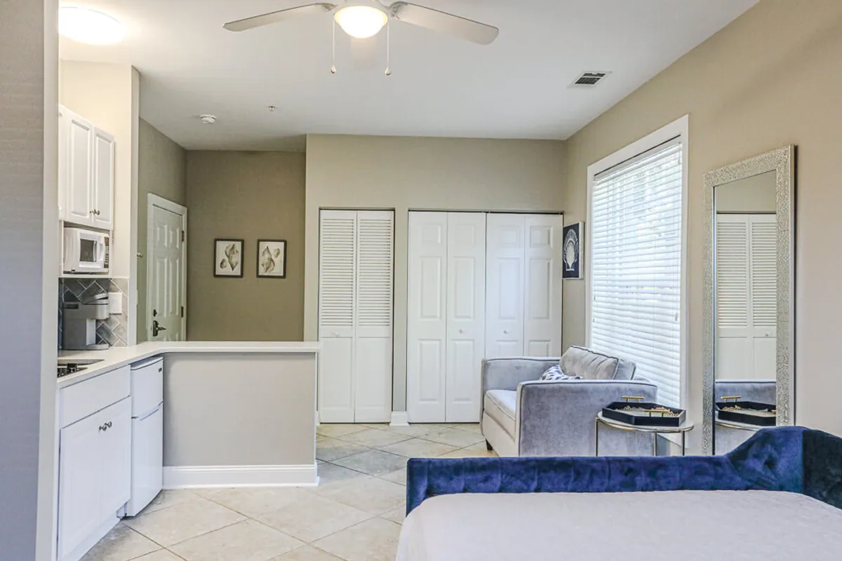 A contemporary studio room with an inviting bed, chic sofa, kitchenette, and stylish mirror, arranged in an open-plan layout. The space is bathed in bright natural light, highlighting the comfortable and modern atmosphere perfect for both relaxation and socializing.